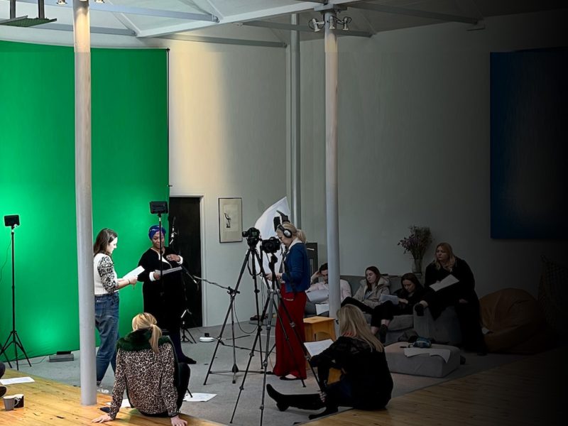 A group of people is filming in a studio with a green screen backdrop. Camera and lighting equipment are set up.