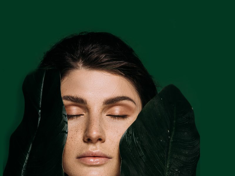 A woman with closed eyes is surrounded by large green leaves against a dark green background.