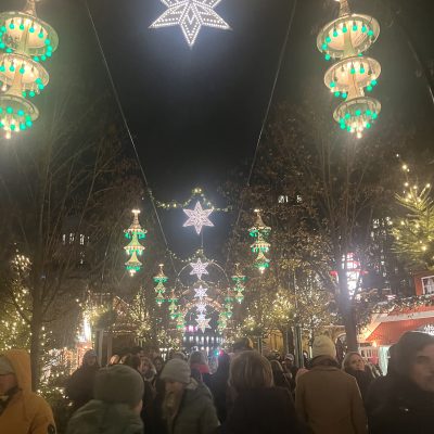 A bustling Christmas market at night with star-shaped and green lanterns hanging above a crowd, surrounded by illuminated trees.