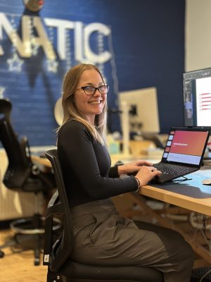 A woman with glasses sits at a desk, smiling, and working on a laptop in a modern office space.