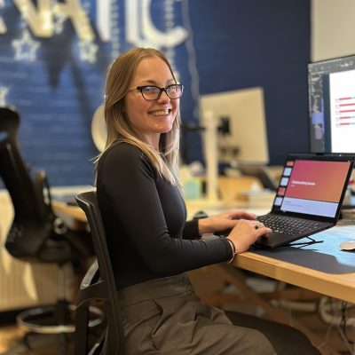 A woman with glasses sits at a desk, smiling, and working on a laptop in a modern office space.