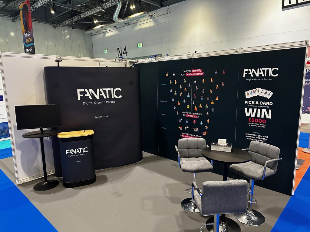 Trade show booth featuring company branding, gray chairs around tables, and promotional signs with a contest offering a €5000 prize.