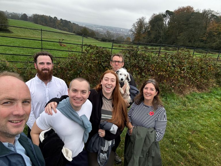 A group of six people and a dog posing happily in a rural setting with rolling green hills and a fence in the background.