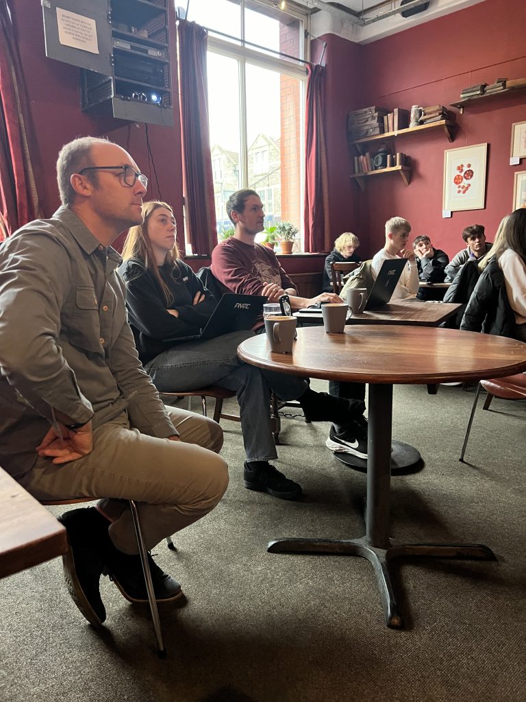 People attentively listening to a presentation in a cozy room with red walls, sitting at wooden tables, some with laptops and coffee.