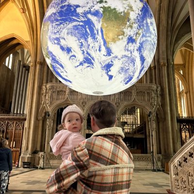 A person holds a baby, gazing at a large Earth model suspended in a cathedral with ornate arches and stonework.