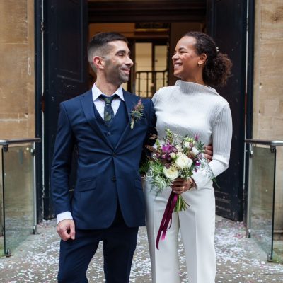 A couple stands arm in arm, smiling at each other. The man is in a navy suit, and the woman in white holds a bouquet of flowers.