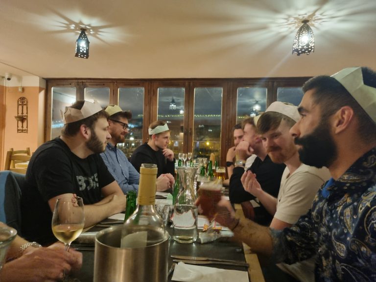 Group of people wearing paper hats, sitting around a dining table with drinks, engaged in conversation, in a warmly lit room.