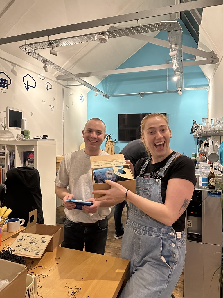 Two smiling people in an office with blue walls. One holds an open box with a mug inside, while the other holds a phone.
