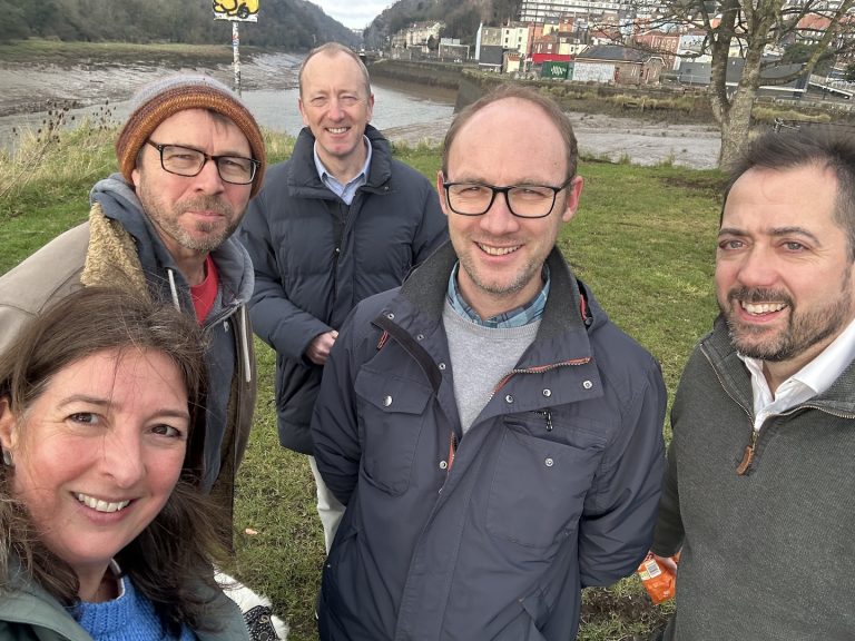 Five people smiling outdoors with a river and hills in the background. They are dressed warmly in jackets, standing on grass.