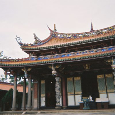 Traditional Chinese temple with ornate, colorful roof tiles, decorative carvings, and stone pillars, set against a cloudy sky.