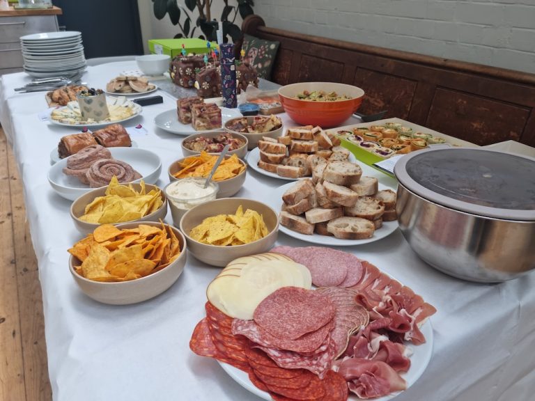 A buffet table with assorted chips, dips, cured meats, sliced bread, pastries, and a large covered bowl on a white tablecloth.