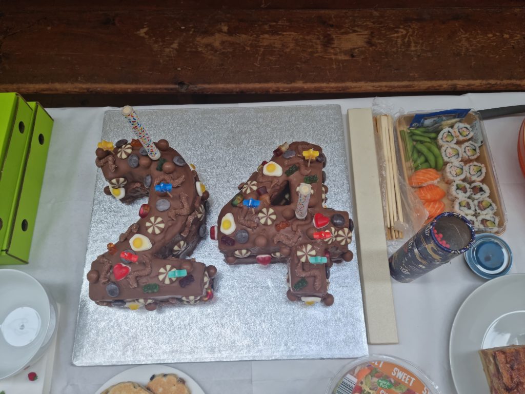 Chocolate cake shaped as the number 24, decorated with candies and chocolates. Sushi and snacks are nearby on the table.