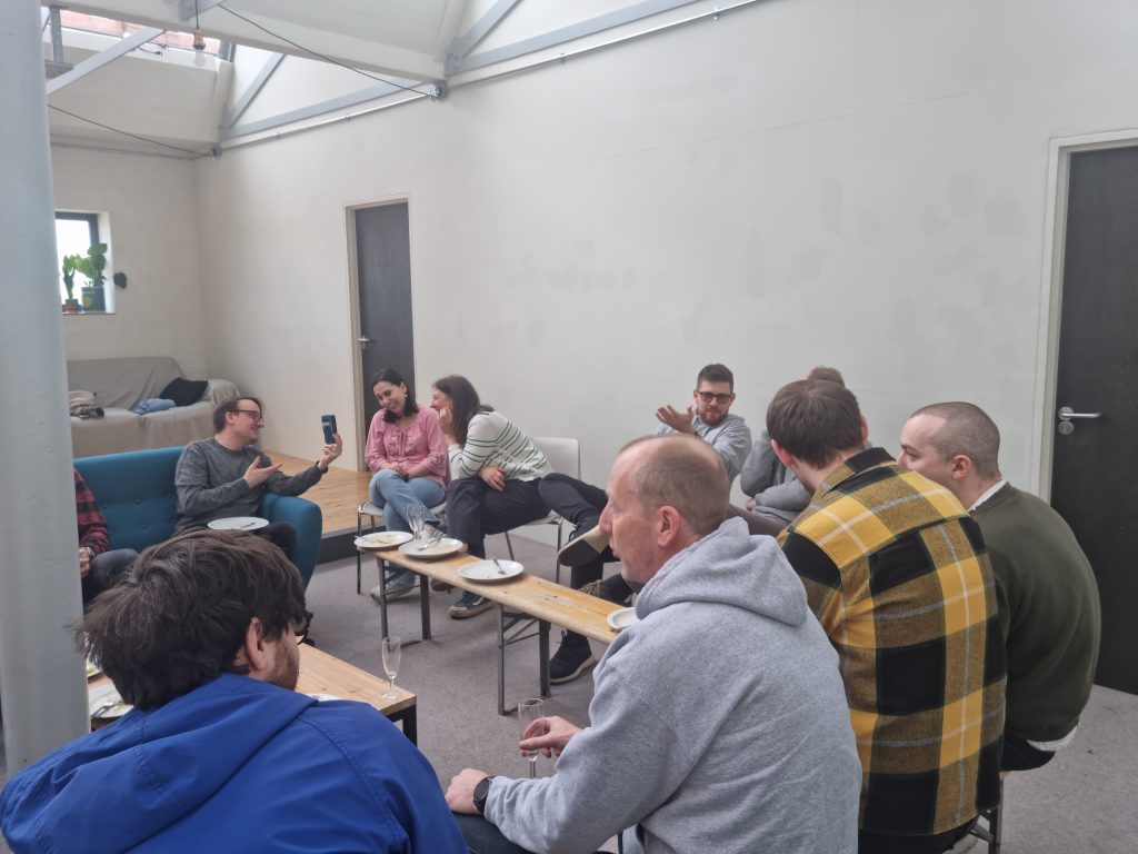 A group of people sitting in a casual setting, talking and looking at a phone. Plates and glasses are on the tables.