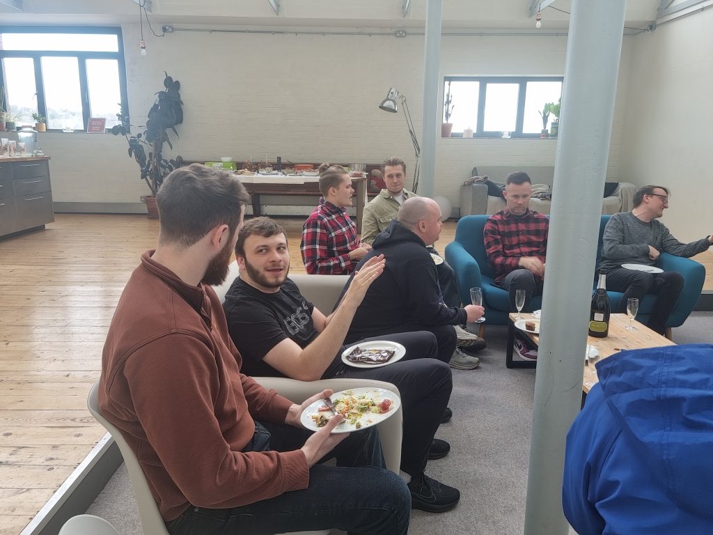 A group of people sitting on couches in a casual setting, holding plates of food and talking. A table with more food is in the background.