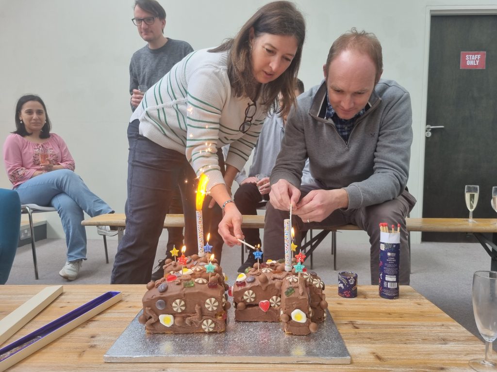 Two people light sparklers on a chocolate cake decorated with colorful candies, with others seated in the background.