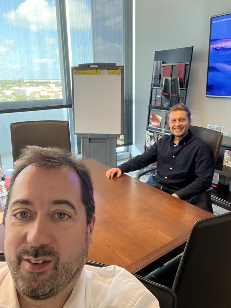 Two people in a meeting room, one taking a selfie. A flip chart, bookshelves, and a large window in the background.
