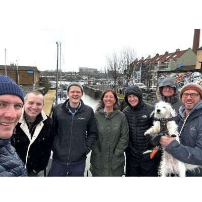 Seven people and a dog pose smiling outdoors by a canal with overcast skies and row houses in the background.