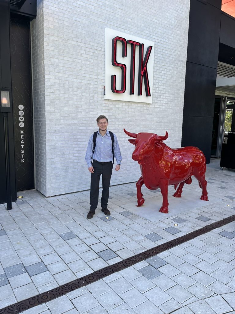 A person stands next to a red bull statue outside a building with a large 
