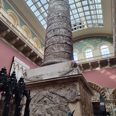 A tall, intricately carved column stands in a spacious room with a glass ceiling and ornate architecture, surrounded by historical artifacts.