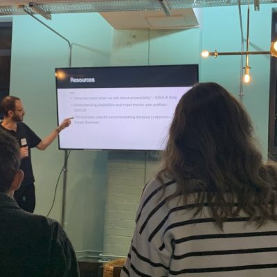 A person gives a presentation on accessibility resources to an audience, with a screen displaying a list of topics behind them.