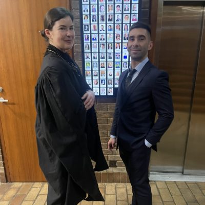 Two people stand in a hallway, one wearing a black robe and the other in a suit. A wall with many small photos is behind them.