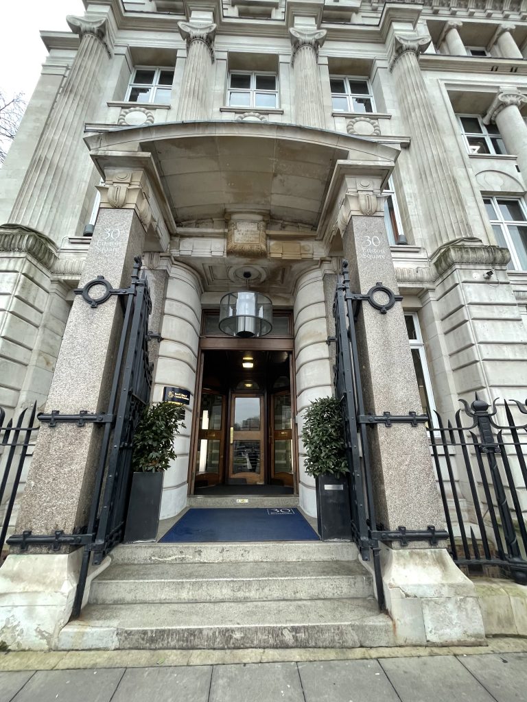 Entrance of a grand building with stone columns, metal gates, and a blue carpet on the steps, flanked by potted plants.