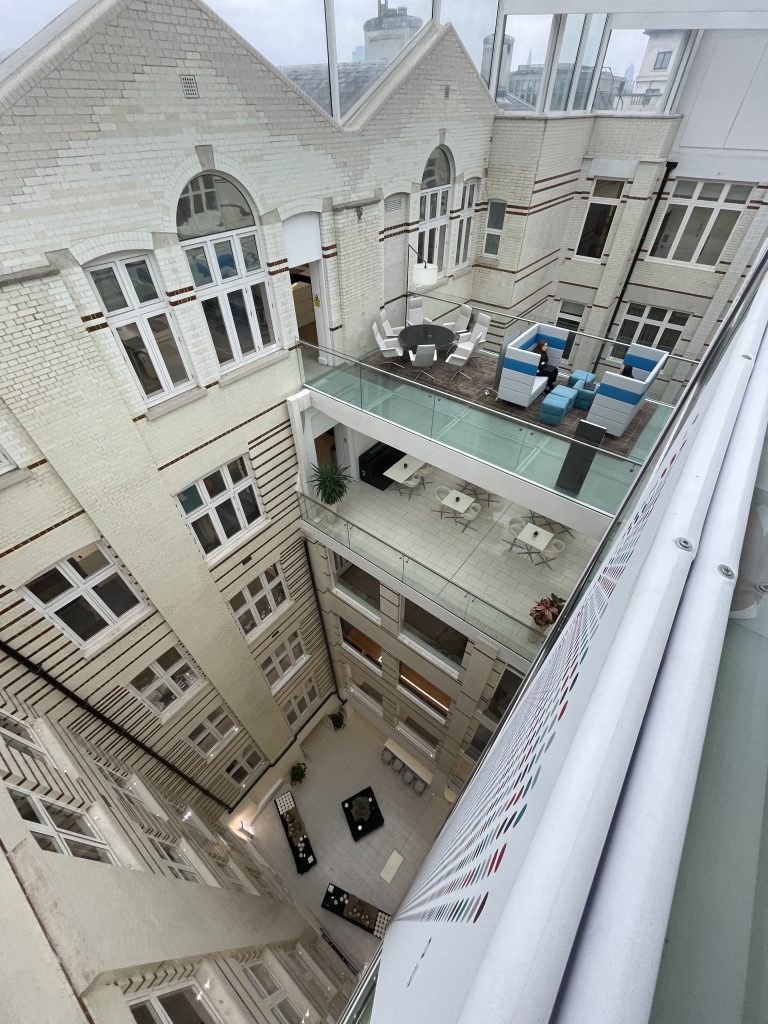 View from high up in a multi-level atrium with balconies, furniture, and white brick walls. Tables and seating areas are visible.