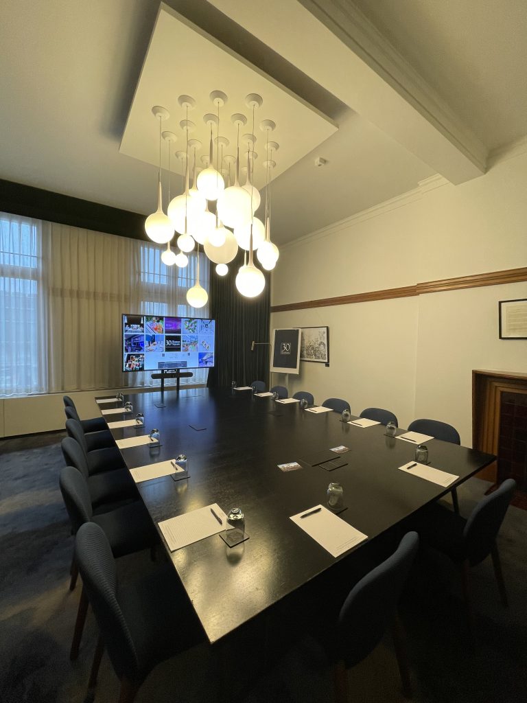 A conference room featuring a long, dark table surrounded by chairs, a modern chandelier, and a screen displaying images.