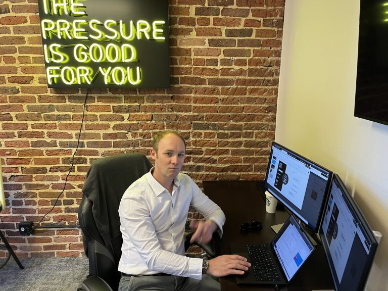 A person sits at a desk with multiple monitors, working on a laptop. A neon sign on the brick wall reads, 