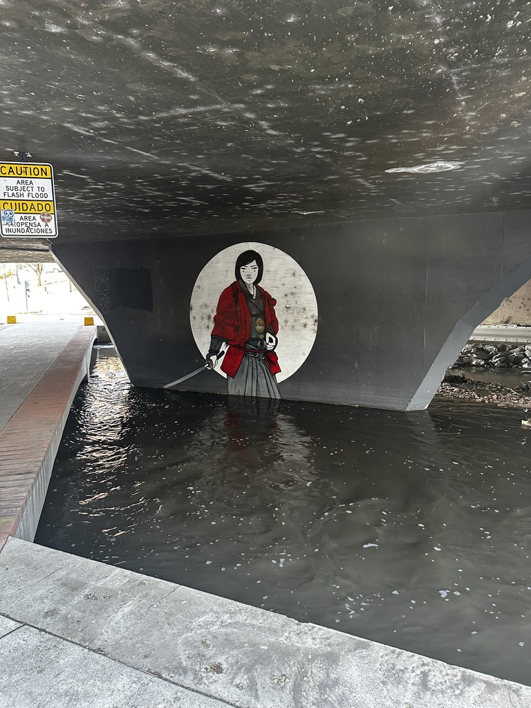 Street art under a bridge shows a person in a red kimono holding a sword. The artwork is partially reflected in the water below.