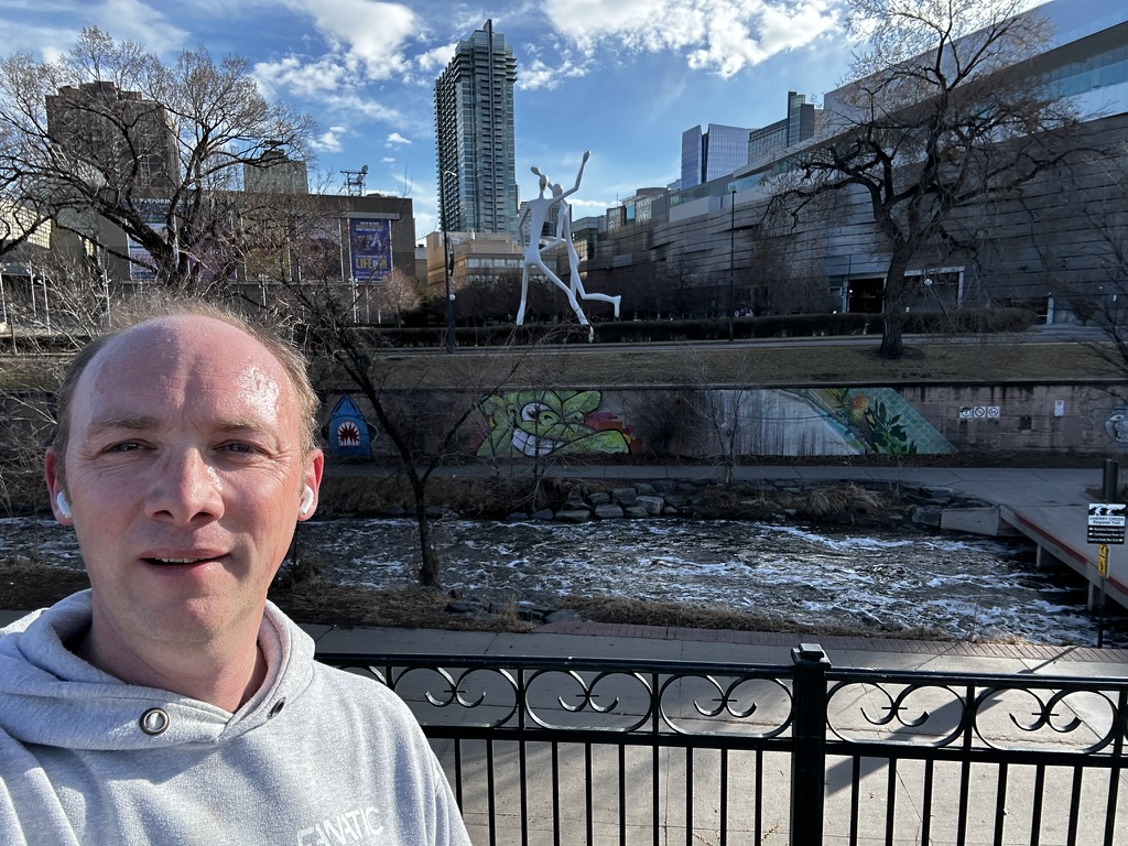 Person in a gray hoodie taking a selfie in a park with a creek, large sculpture, and city buildings in the background.
