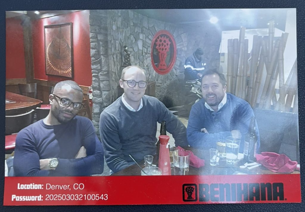 Three people seated at a restaurant table, smiling. They are surrounded by drinks and condiments, with a red-and-stone decor theme.
