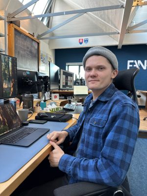 A person in a beanie and blue plaid shirt sits at a desk with multiple monitors. The workspace has a casual, creative vibe.