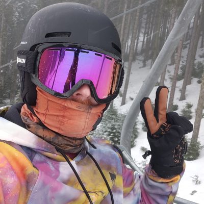 Person wearing a ski helmet and goggles flashes a peace sign while on a ski lift. Snowy trees are visible in the background.