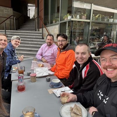 Six people are smiling and seated around an outdoor café table with drinks and pastries. There's a staircase in the background.