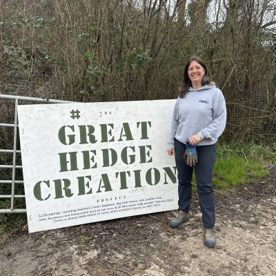A woman in outdoor clothing stands beside a sign reading 