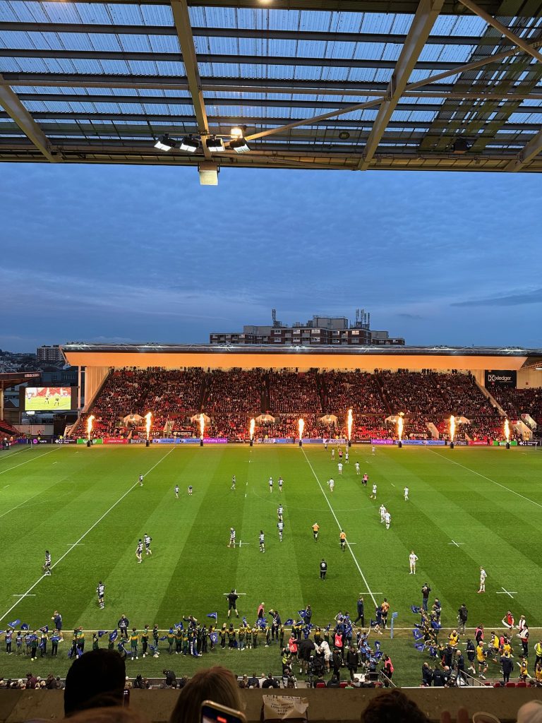A rugby match is taking place in a stadium with a large crowd. Flames are shooting up along the sidelines, and dusk is approaching.