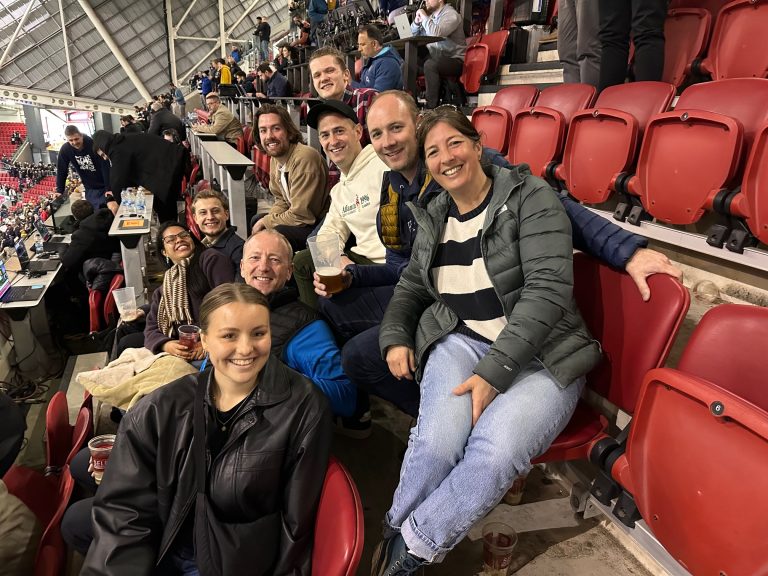 A group of people smiling and sitting in a stadium's red seats, holding drinks and wearing casual attire.