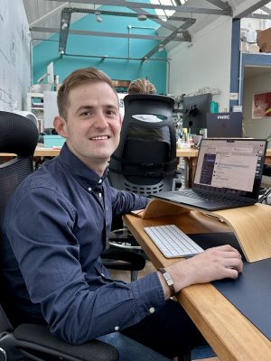 A person in a blue shirt sits at a desk with a laptop and a wooden laptop stand, smiling towards the camera in a modern office.