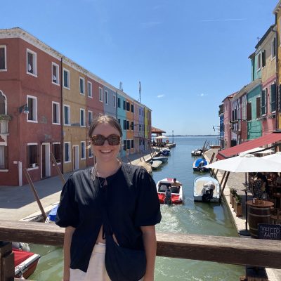 A person in sunglasses stands smiling on a bridge in front of colorful buildings lining a canal, with boats and water visible under a clear blue sky.