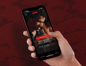 A hand holds a smartphone displaying a restaurant app screen showing a smiling person. The background features a dark, decorative pattern.