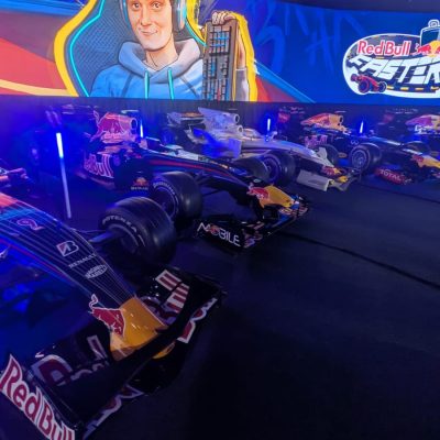 Row of Red Bull racing cars displayed indoors with colorful lighting and a large screen featuring a graphic of a person and the logo.