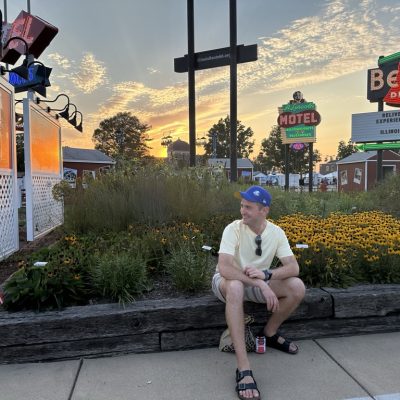 Man in a yellow shirt and blue cap sitting on a wooden ledge with yellow flowers, surrounded by vintage signs and a sunset backdrop.
