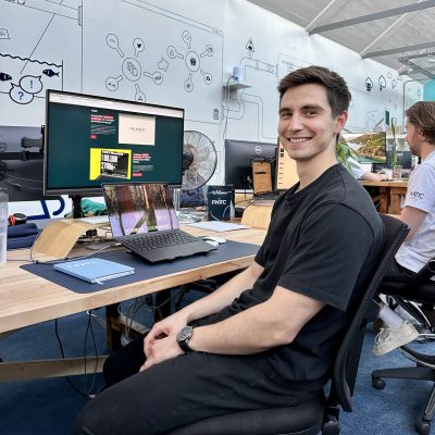 A person in a black shirt sits at a desk with a laptop and monitor, smiling at the camera in a modern, creative office space.
