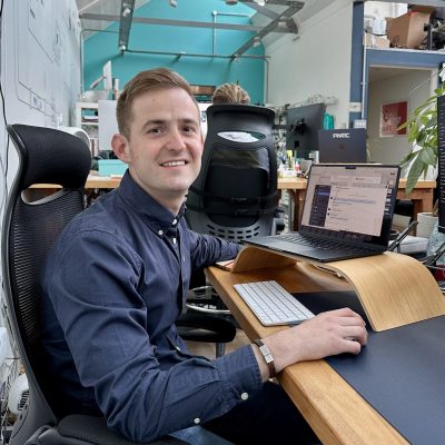 A person in a blue shirt smiles at a desk with a laptop, keyboard, and wooden stand in an office environment.