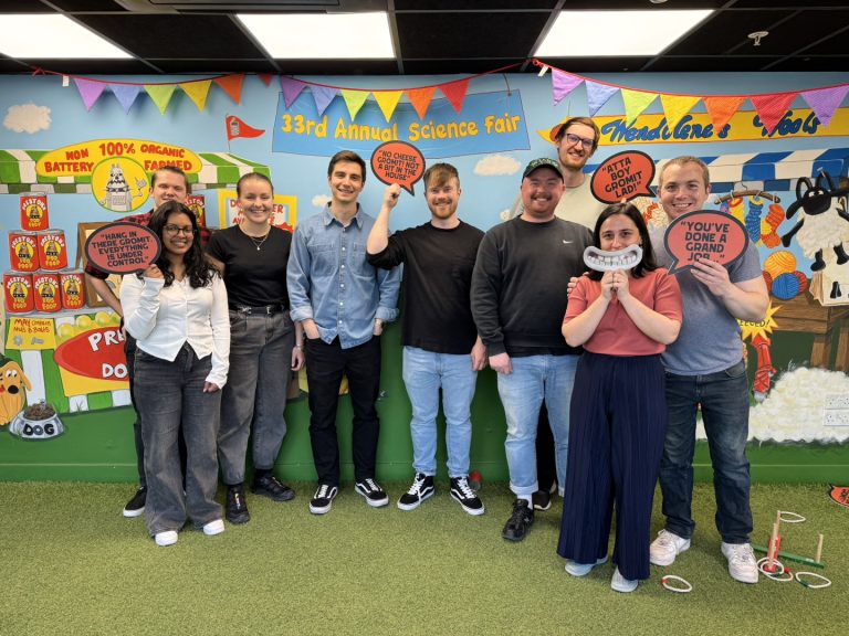 A group of eight people smiling and holding props in front of a colorful science fair backdrop with banners and decorations.