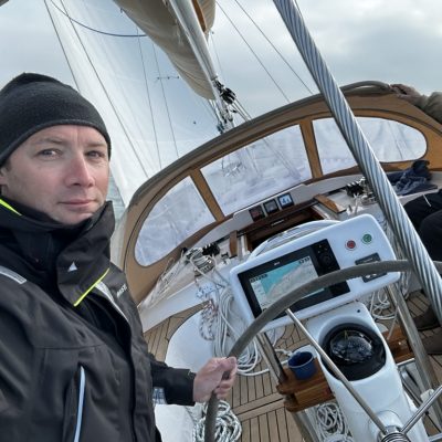Person in a black jacket and beanie steering a sailboat, with navigational instruments visible and a cloudy sky in the background.