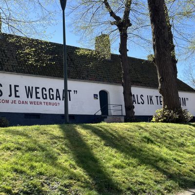 A white brick building with a tiled roof and Dutch text painted on the side, surrounded by trees and grass under a clear blue sky.