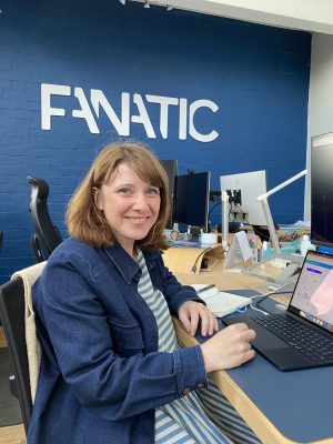 A person smiling at the camera while sitting at a desk with a laptop. The word 