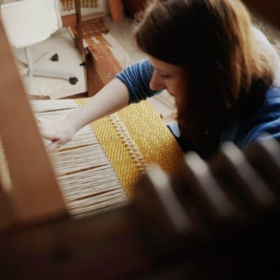 A woman weaving a yellow patterned fabric on a wooden loom, focusing intently on her work.